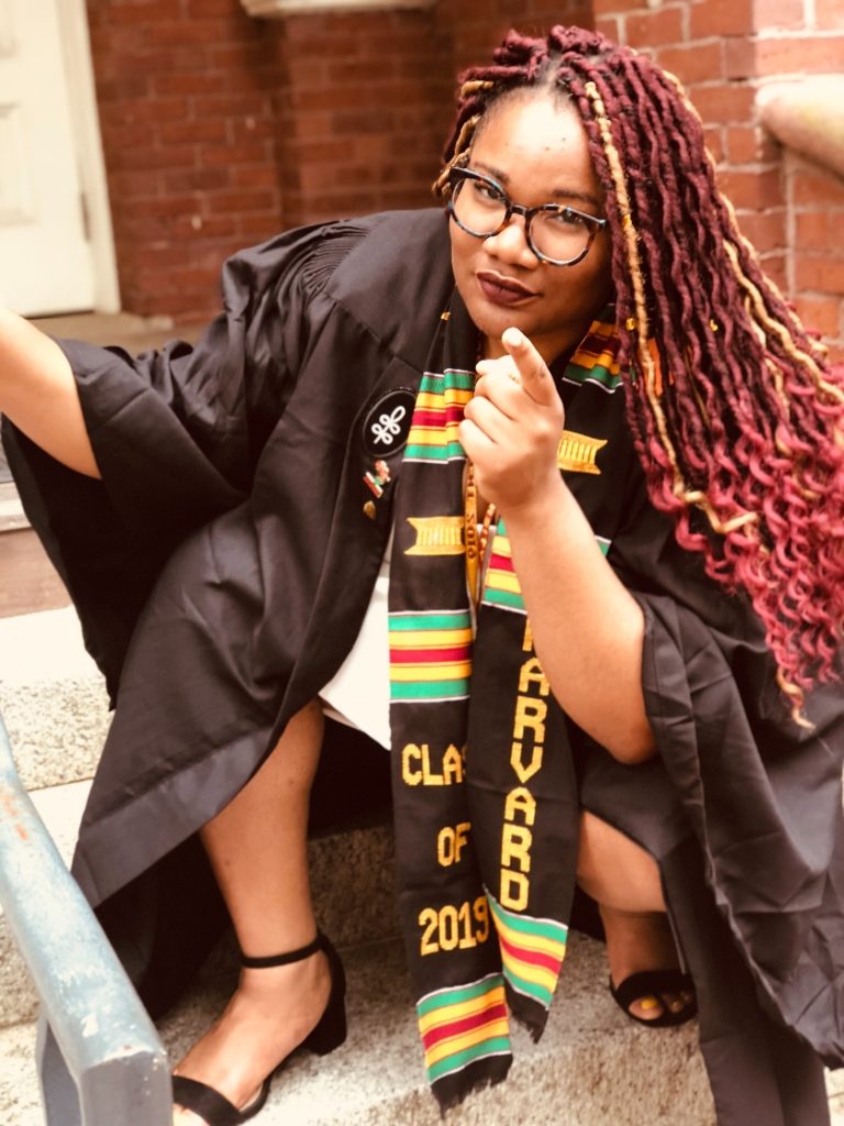 Drey pointing at camera during her Harvard graduation while wearing her graduation robe in 2019.